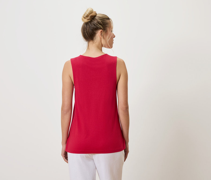 Widok z tyłu kobiety z kokiem na głowie, ubranej w czerwony top na ramiączkach i białe spodnie.