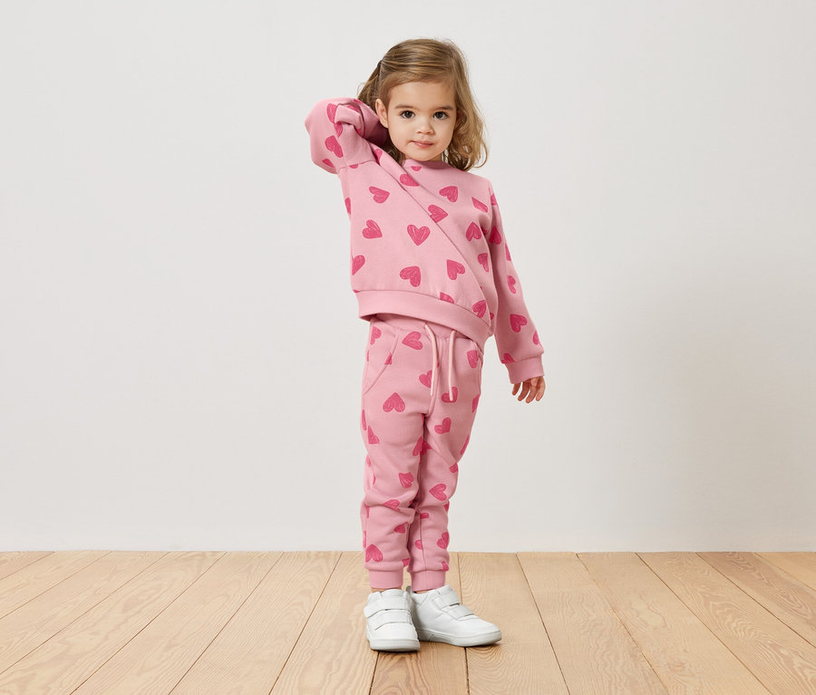 Dziewczynka z blond włosami ma na sobie różowy sweter i spodnie w serduszka oraz białe tenisówki.