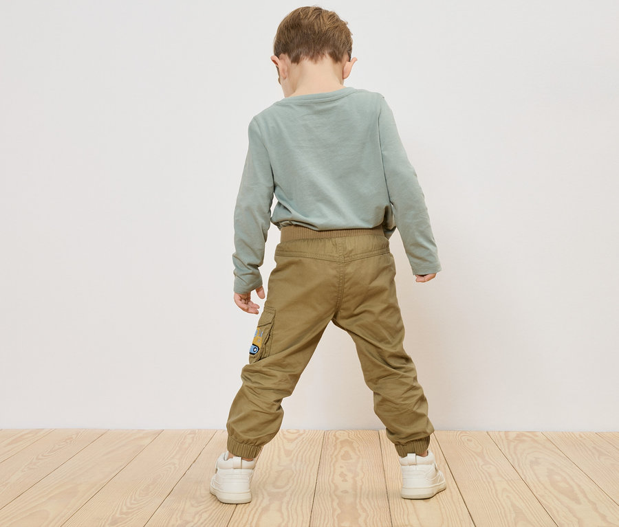 Dziecko stoi tyłem do kamery, ma na sobie zieloną koszulkę z długim rękawem, khaki spodnie i białe trampki.