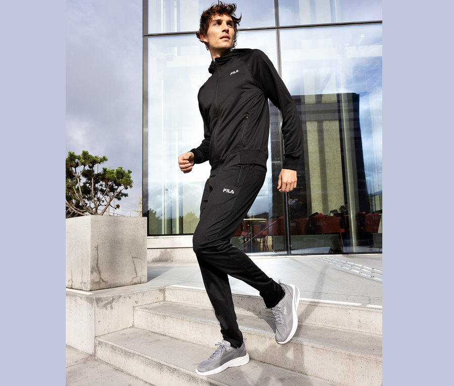 Mężczyzna w czarnym dresie Fila i szarych butach sportowych schodzi po betonowych schodach.