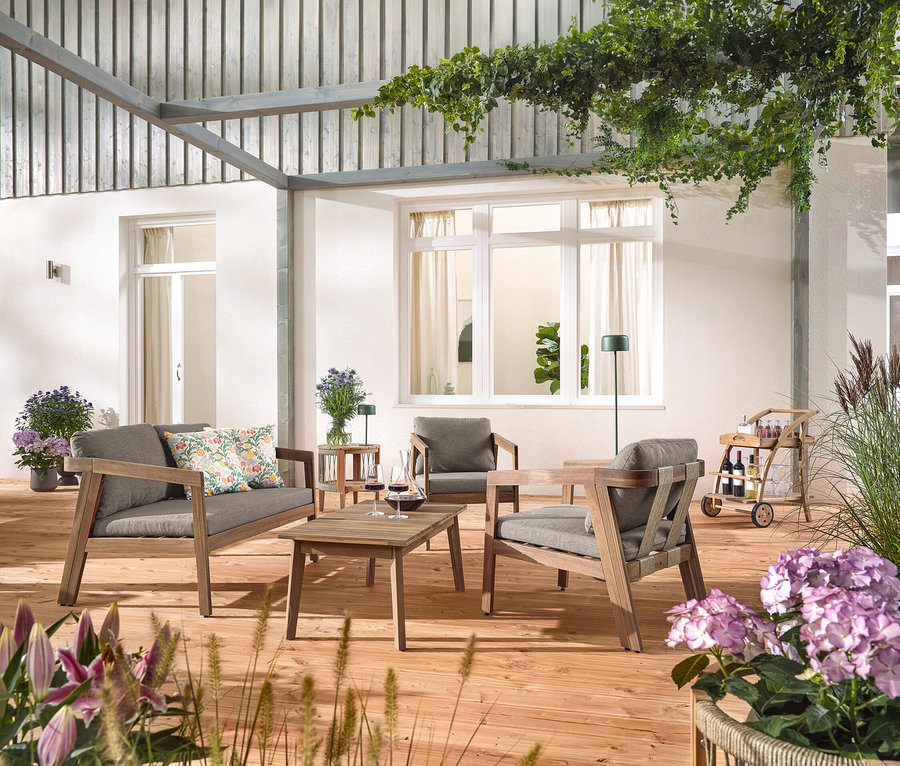 Na drewnianym tarasie stoi 4-częściowy zestaw wypoczynkowy do ogrodu z plecionki tekstylnej, z 2 poduszkami ogrodowymi we wzory kwiatowe, wózek barowy z odłączaną tacą i stolik pomocniczy do ogrodu z odłączanym blatem, Ø ok. 50 cm. Widoczna jest również solarna lampa stołowa z akumulatorem, dżungla zielona i solarna lampa podłogowa z akumulatorem, dżungla zielona.