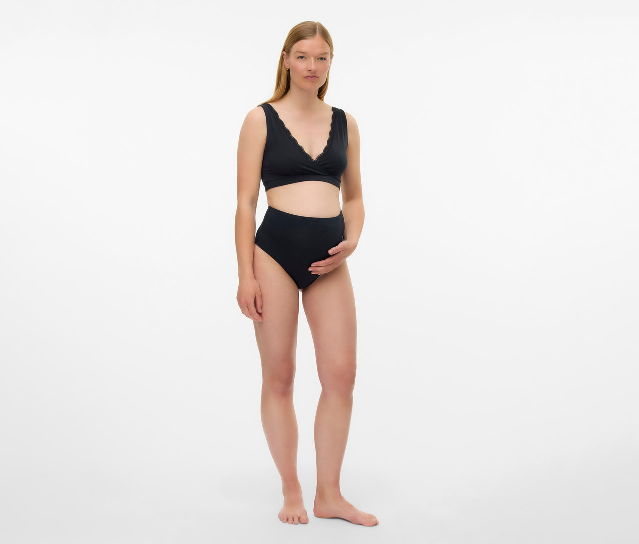 Kobieta w ciąży ma na sobie czarny biustonosz ciążowy MAMALICIOUS „MLHELENA” z 2 sztuk i majtki z wysokim stanem na białym tle.