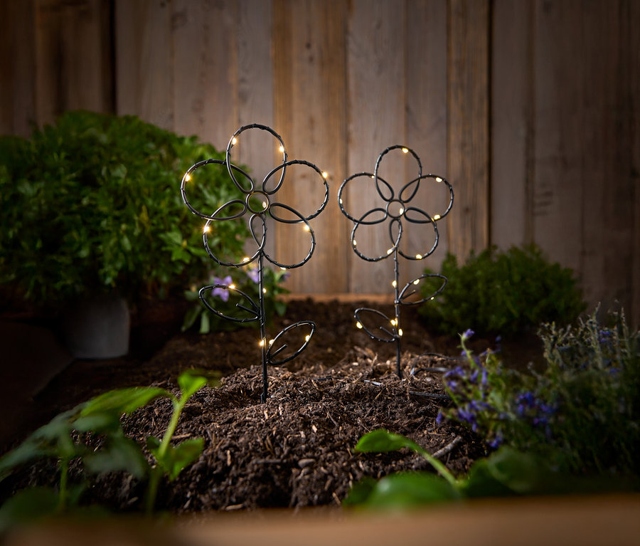 Dwa czarne, ogrodowe słupki w kształcie kwiatów ze światełkami LED stoją w ziemi na tle drewnianej ściany.