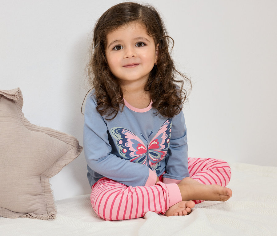 Dziewczynka z kręconymi włosami siedzi na łóżku, ubrana w niebieską piżamę z motylem i różowe spodnie w paski.