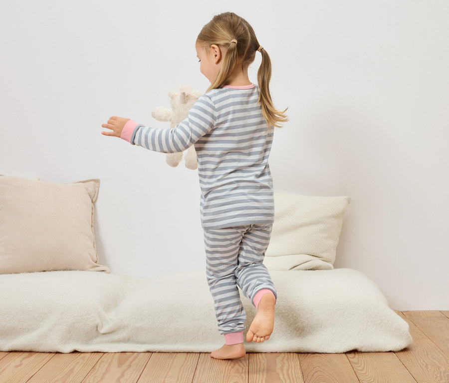 Dziewczynka w szaro-białej piżamie w paski trzyma pluszowego misia na kanapie.