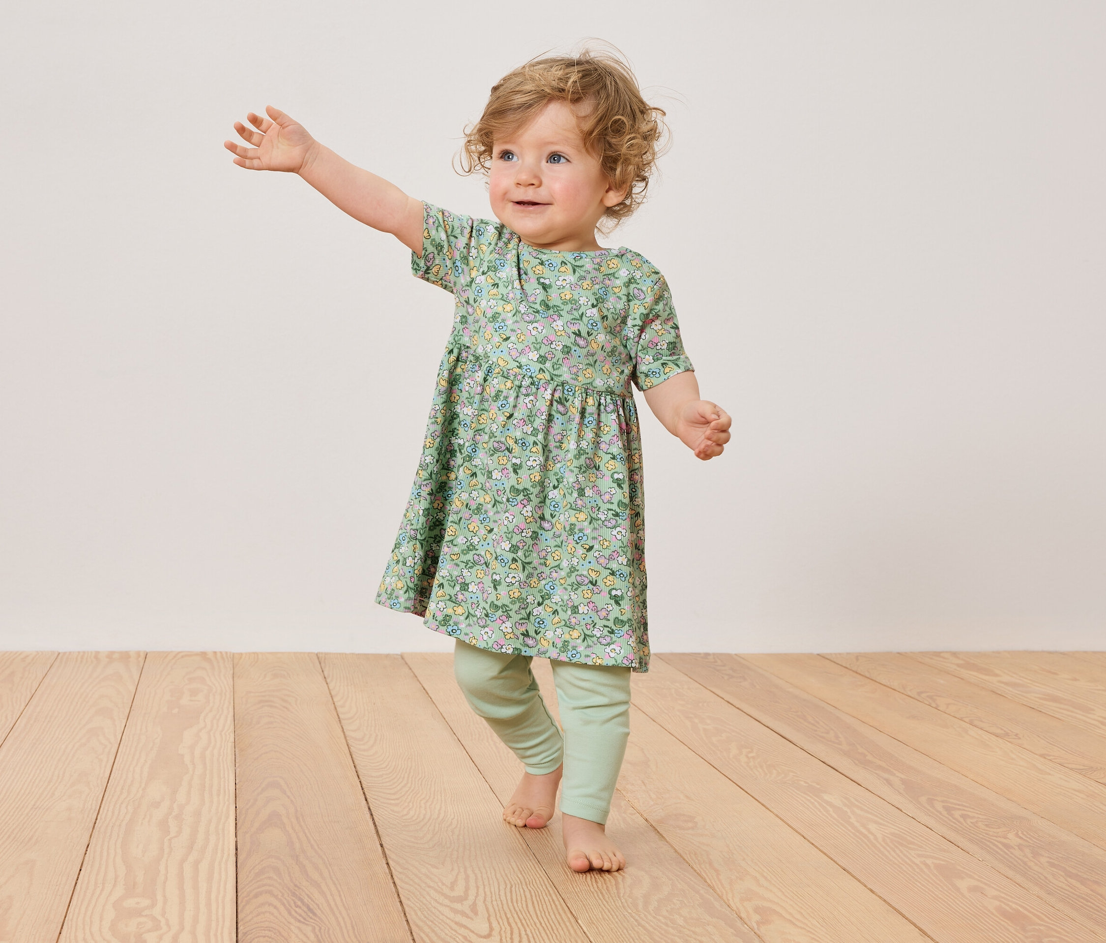 Małe dziecko z blond lokami ma na sobie kwiecistą sukienkę i zielone legginsy, stojąc na drewnianej podłodze.