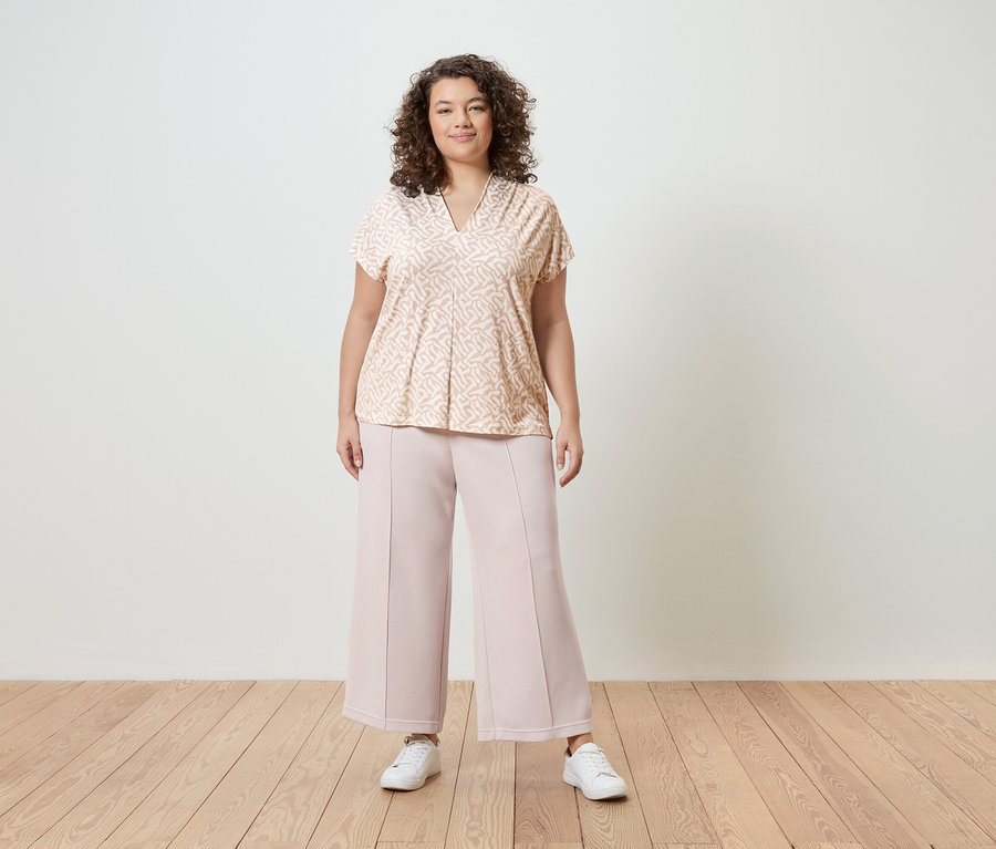 Kobieta o kręconych włosach ma na sobie bluzkę w jasnoróżowe wzory, różowe spodnie typu culottes i białe trampki na tle białej ściany.