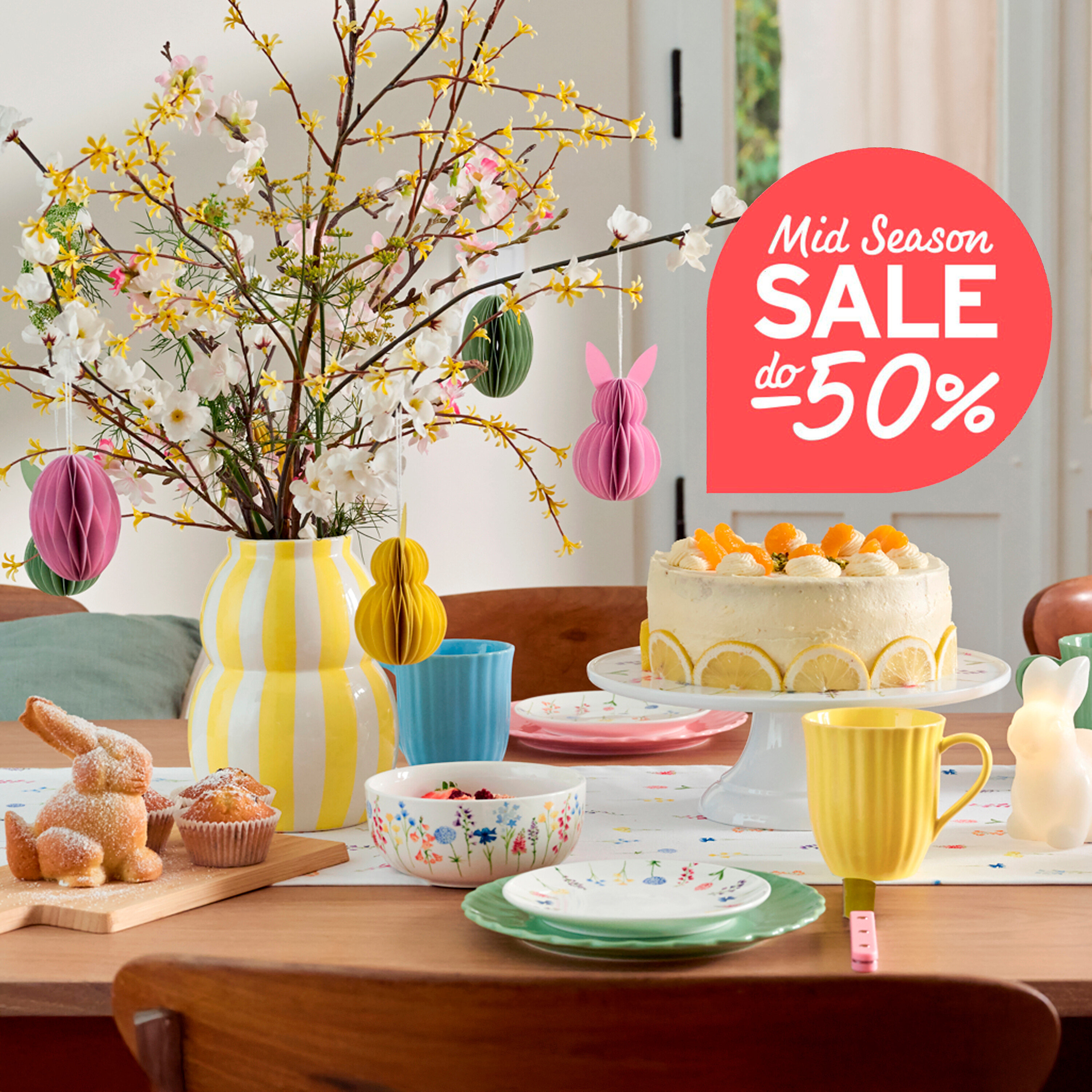 Na stole stoi wazon z kwitnącymi gałązkami i kolorowymi pisankami, naczynia, ciasto i chleb w kształcie zajączka.