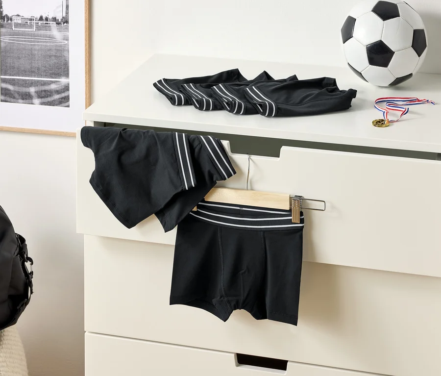 Na komodzie leżą czarne bokserki z białymi paskami, piłka do piłki nożnej i medal. Na wieszaku wisi para spodenek sportowych.