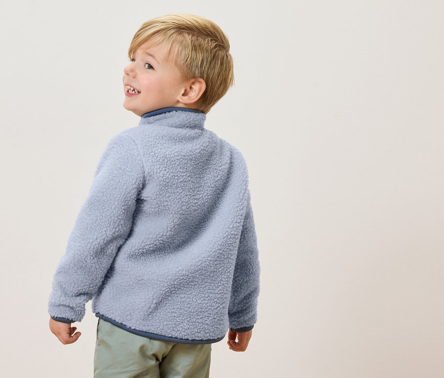 Mały chłopiec z blond włosami ma na sobie jasnoniebieską bluzę polarową i jasnozielone spodnie, patrząc w bok.