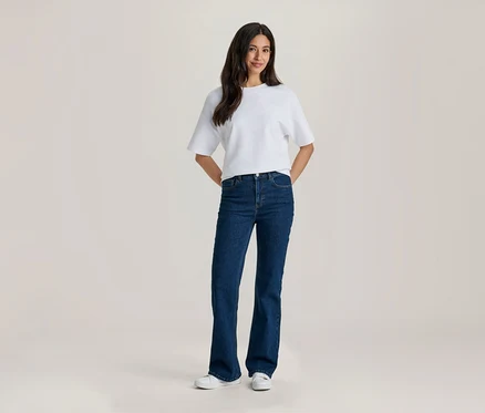 Uśmiechnięta kobieta z długimi ciemnymi włosami stoi w białym T-shircie, granatowych dżinsach dzwonach i białych sneakersach, z rękami za plecami.