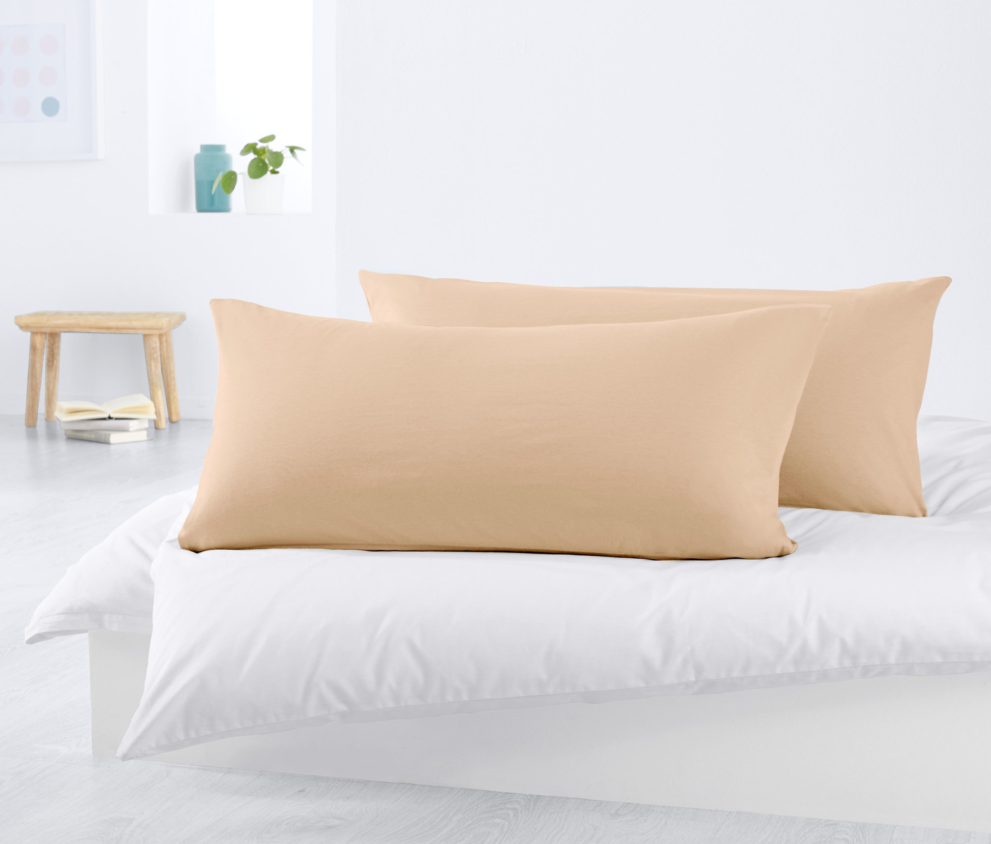 Dwie beżowe dżersejowe poszewki na poduszki leżą na białym łóżku.