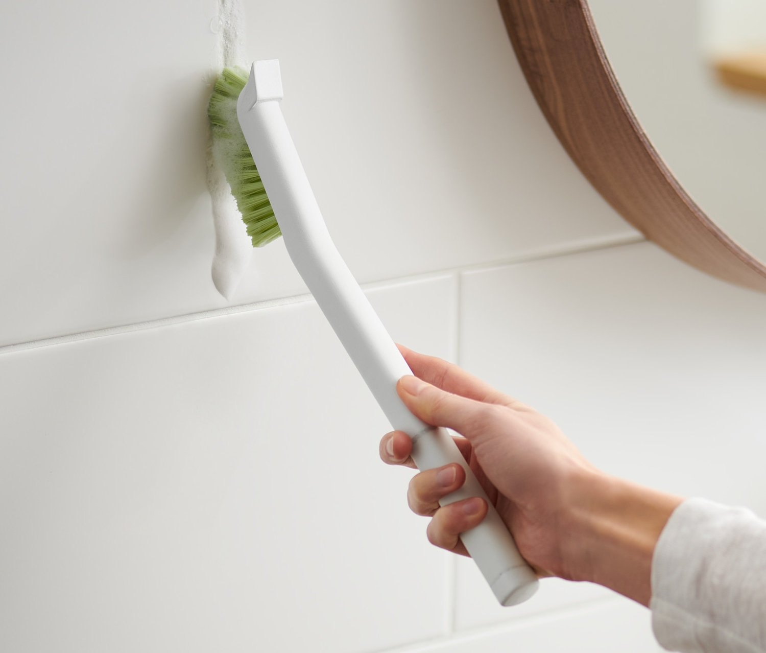 Kobieta czyści fugi między białymi płytkami długą szczotką do fug.