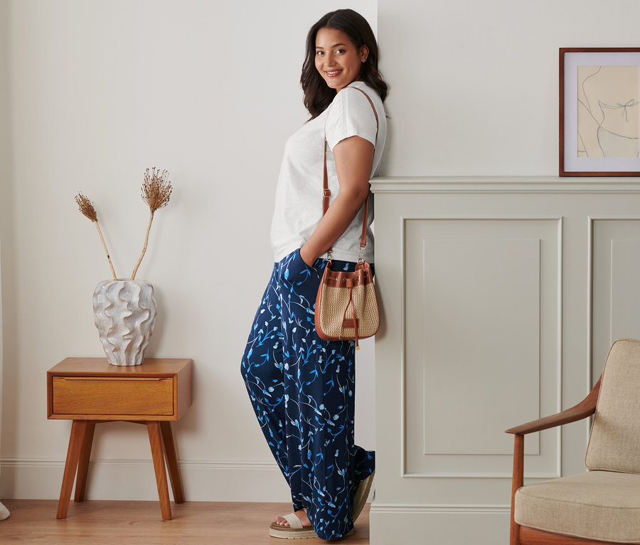 Kobieta oparta o ścianę pozuje w spodniach palazzo, białej bluzce i skórzanych sandałach na platformie. Przez ramię przewieszona ma torbę-worek.