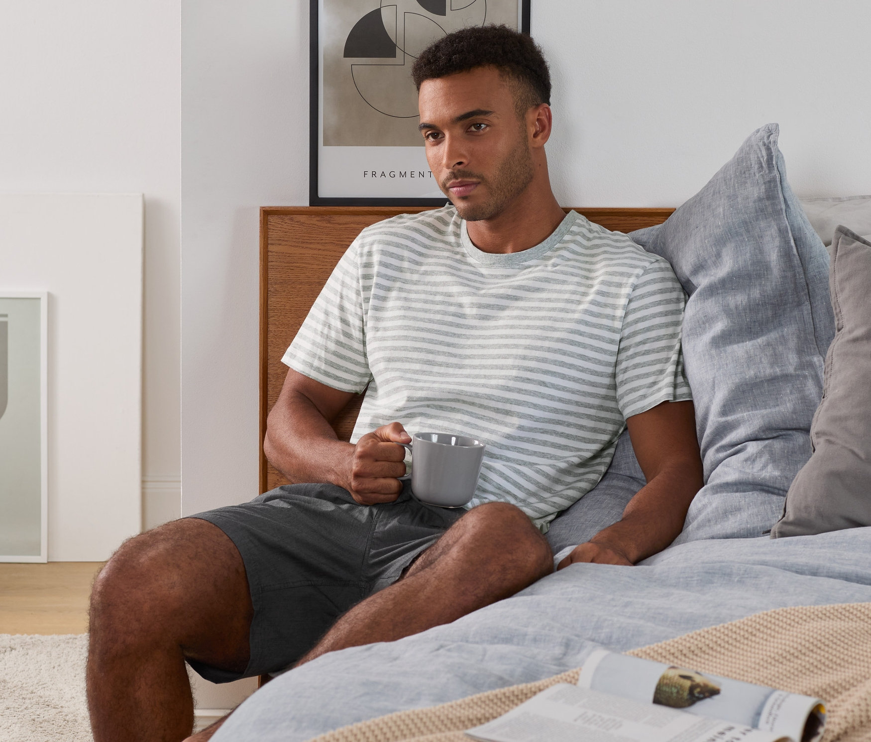 Mężczyzna siedzi na łóżku i trzyma kubek. Ma na sobie piżamę z koszulką i krótkimi spodenkami z tkaniny Chambray.