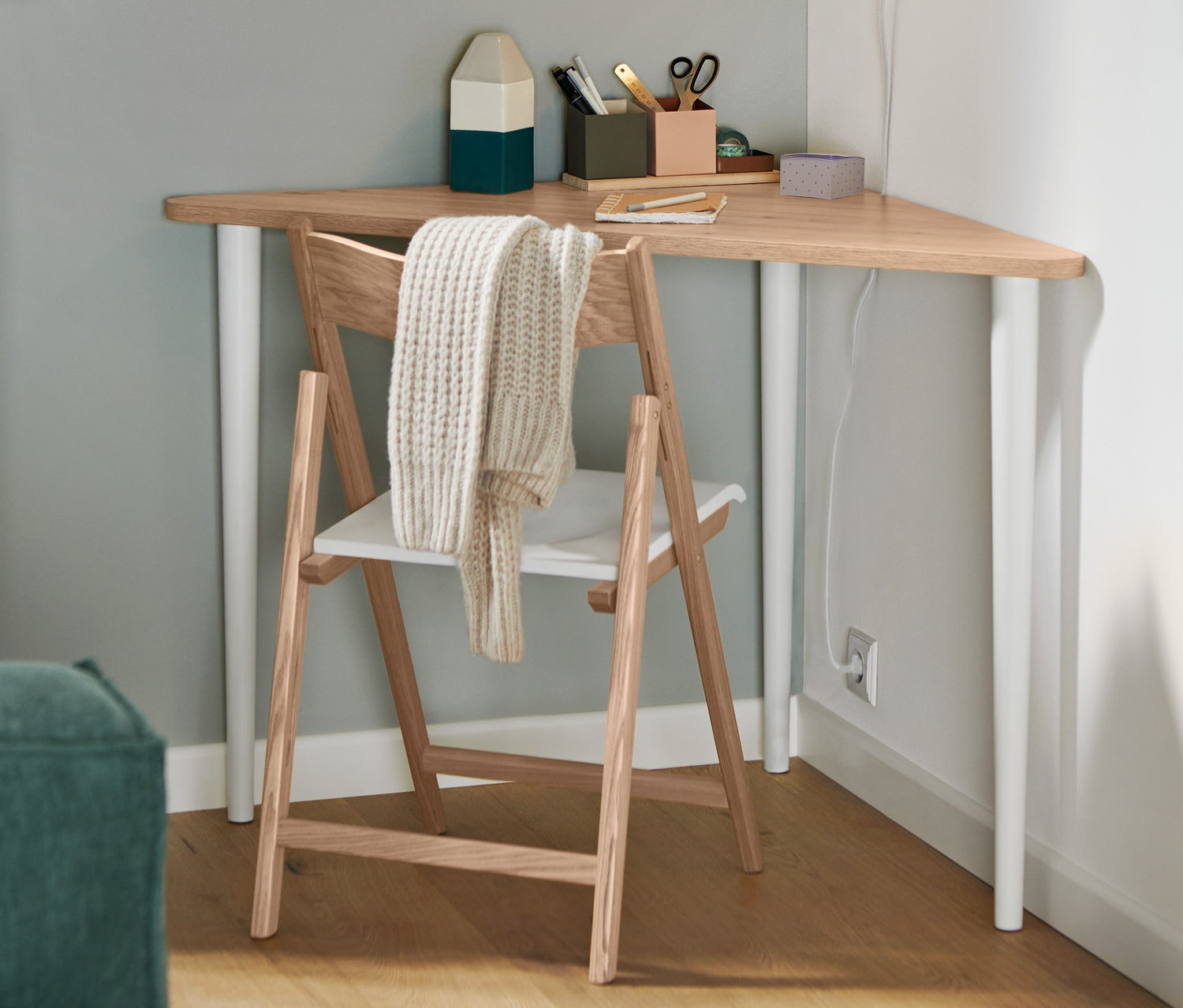 Składane krzesło z drewna jesionowego z certyfikatem FSC® stoi przy trójkątnym stole. Na ścianie wisi biała metalowa lampa ścienna.