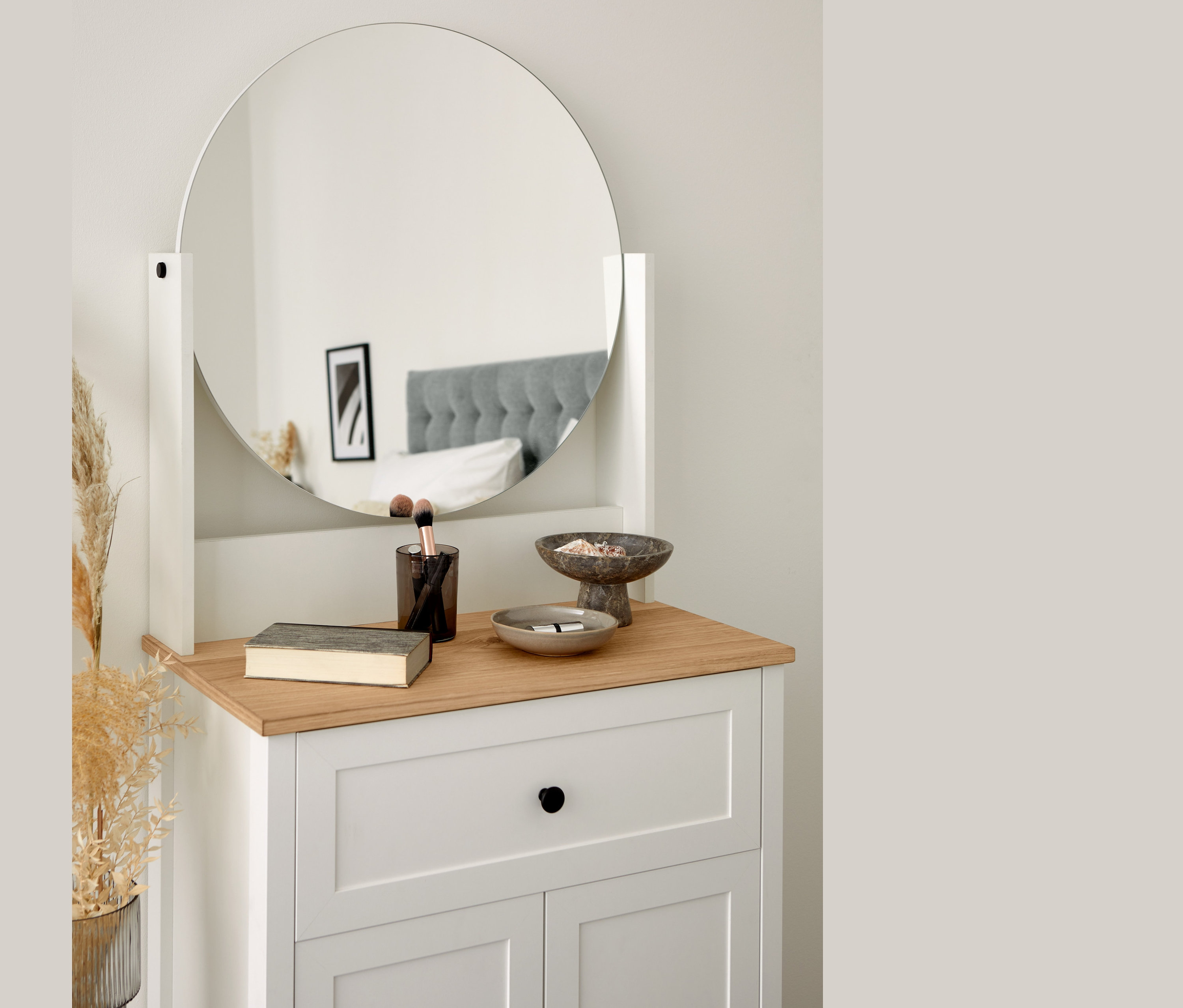 Toaletka z okrągłym lustrem, książką, pędzlami i dekoracyjnymi miseczkami. W lustrze odbija się łóżko.