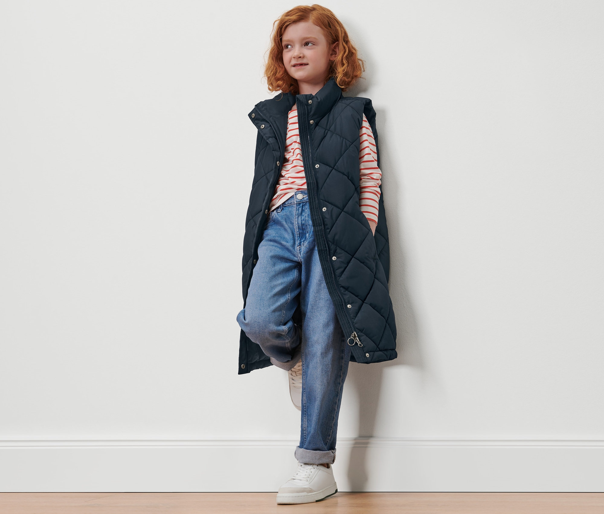 Ruda dziewczyna opiera się o ścianę, ubrana w długą, pikowaną kamizelkę dziecięcą.