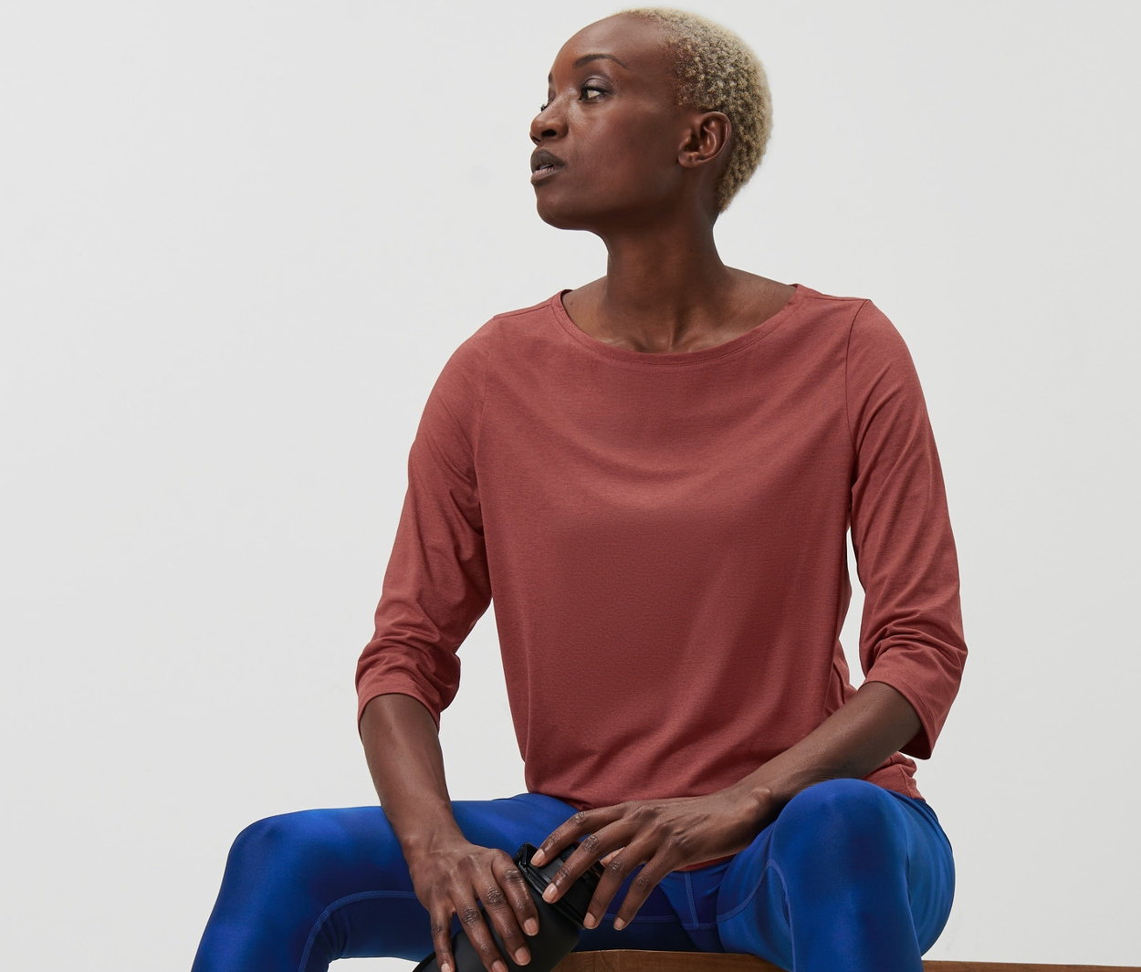 Kobieta siedzi na ławce i trzyma butelkę z wodą. Ma na sobie koszulkę funkcyjną z rękawem o długości 3/4 w kolorze terakoty.