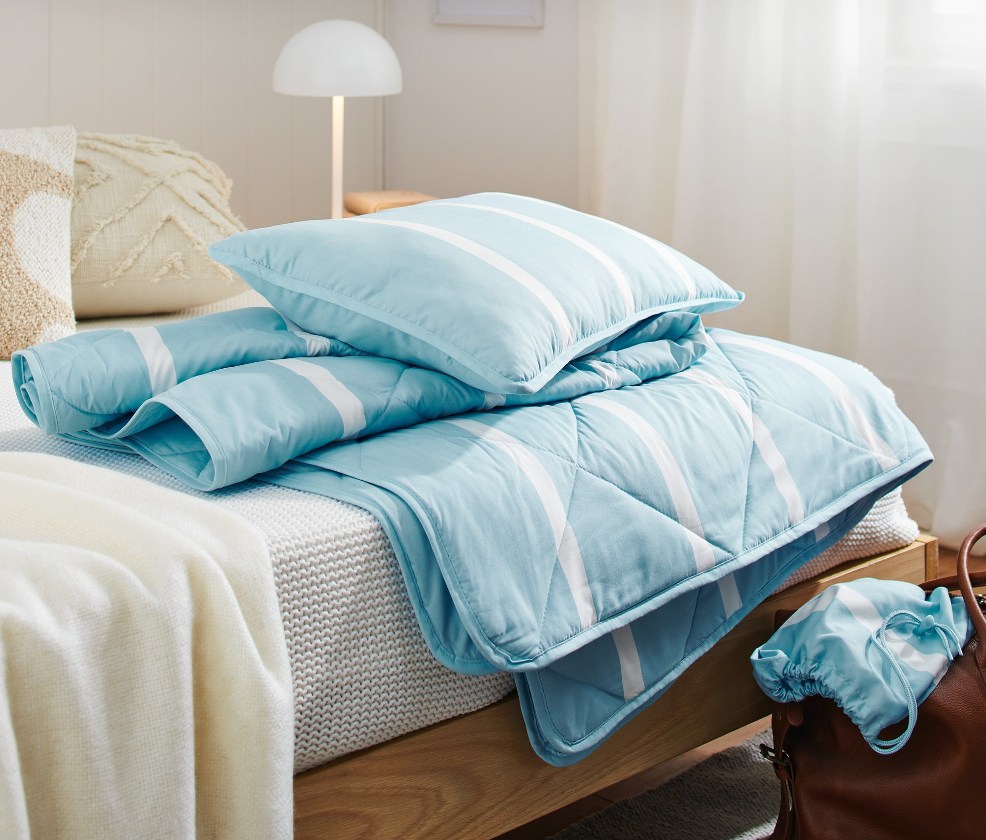 Na łóżku leży turystyczny komplet pościeli w kolorze jasnoniebieskim w białe paski oraz brązowa torba podróżna.