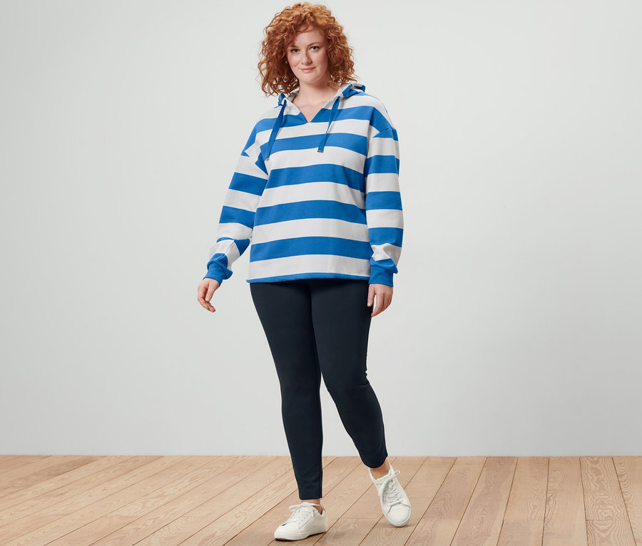 Ruda kobieta pozuje w bluzie z kapturem w paski i czarnych, grubych legginsach.