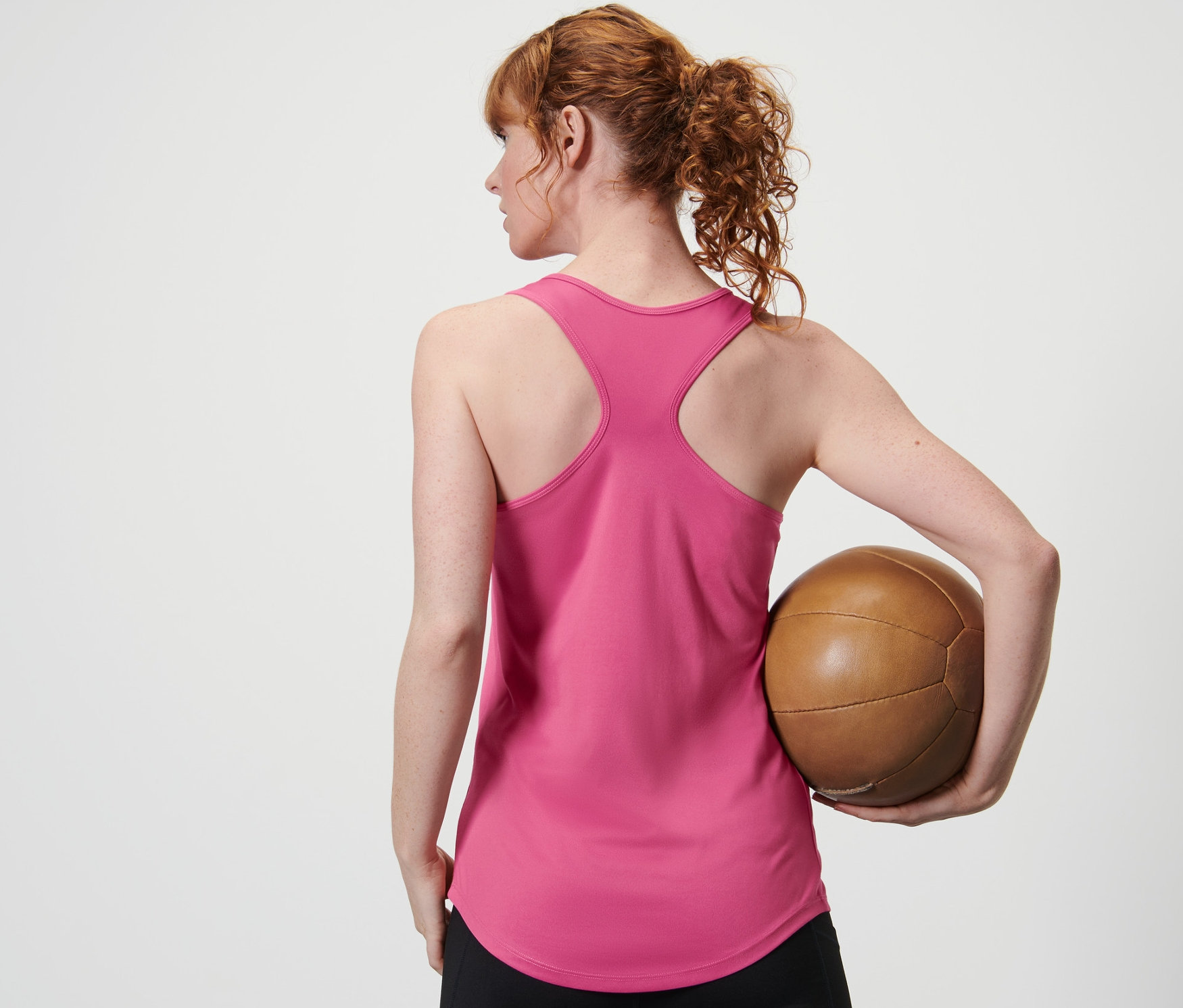 Kobieta trzyma od tyłu brązową piłkę w różowym topie funkcyjnym z dwuczęściowego zestawu i czarnych legginsach.