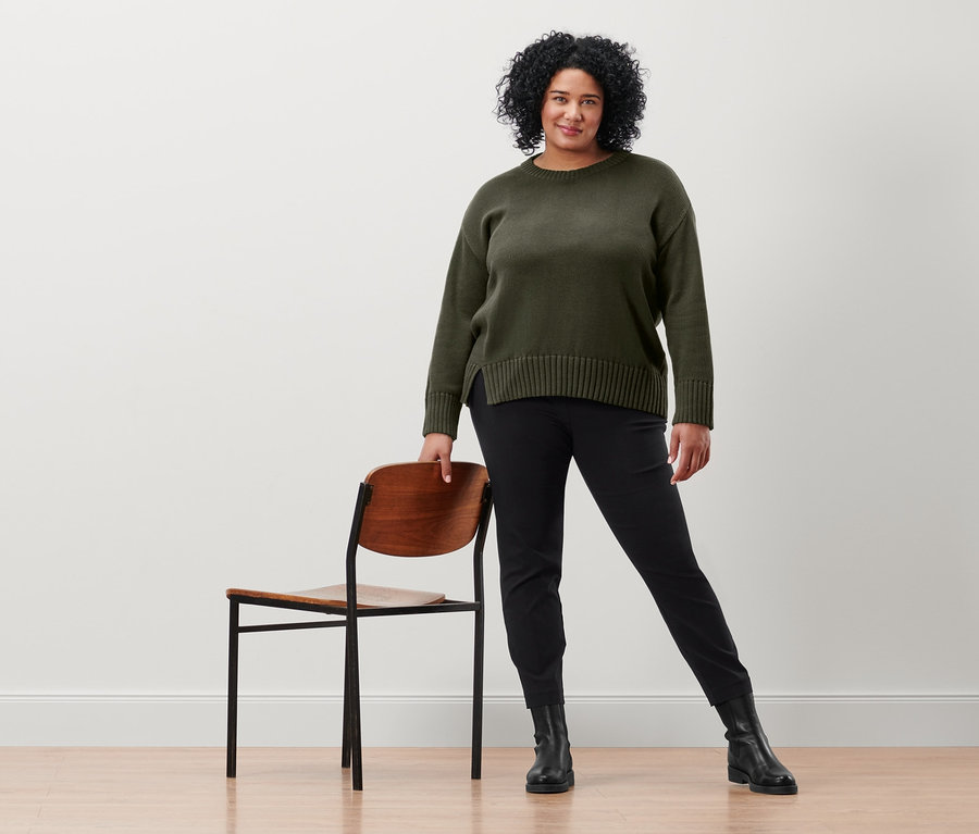 Kobieta pozuje obok krzesła, ubrana w czarne spodnie stretchowe i sweter z dzianiny z rozcięciem u dołu.