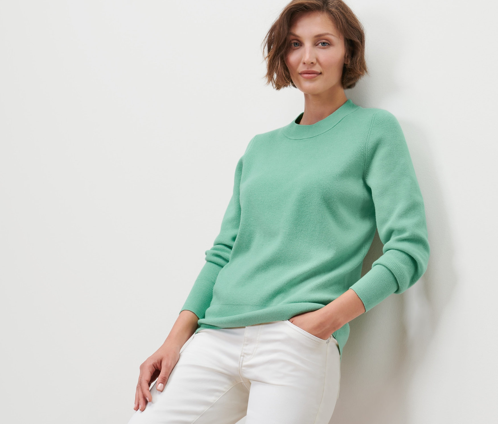 Kobieta oparta o ścianę ma na sobie sweter z dzianiny i białe dżinsy.