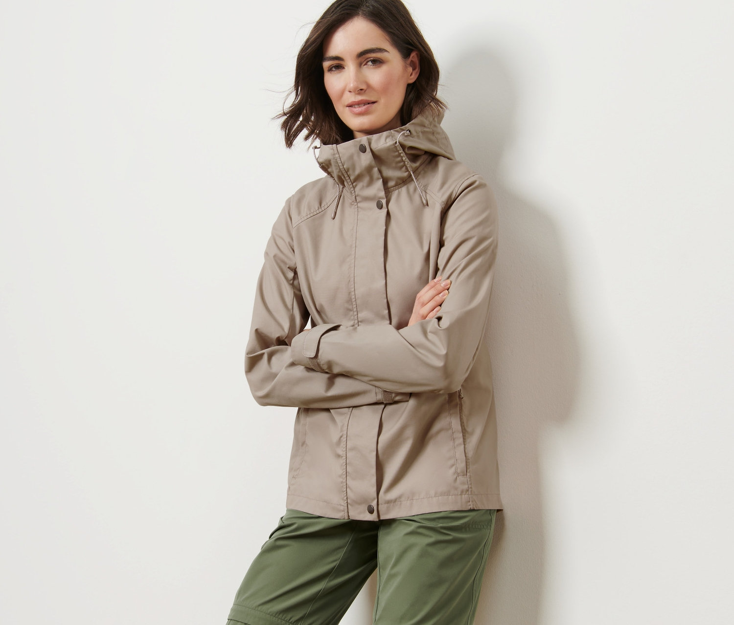 Kobieta pozuje w beżowej kurtce na każdą pogodę i zielonych spodniach.