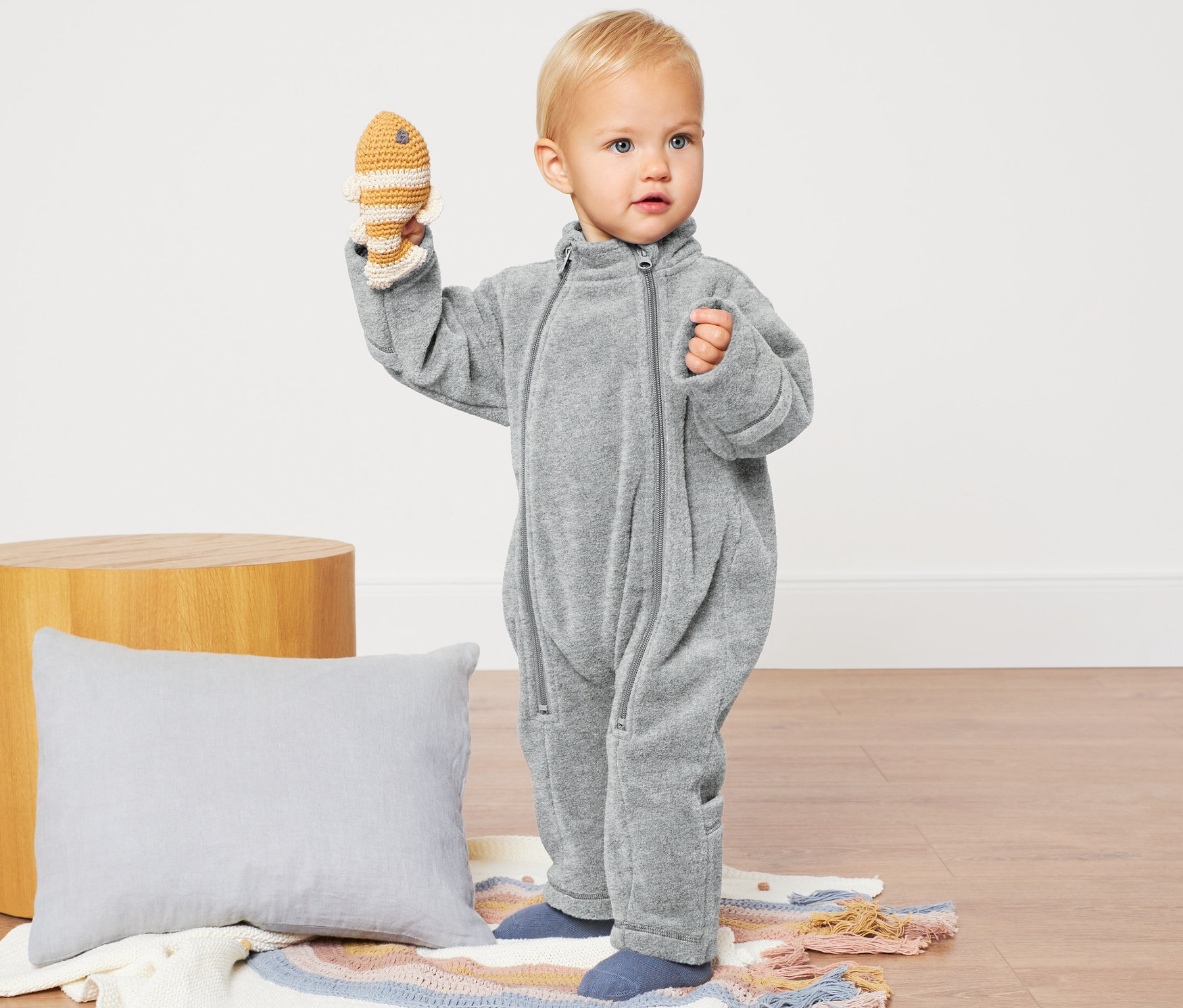 Niemowlę trzyma szydełkowaną rybę, ubrane w szary pajacyk niemowlęcy z polaru.