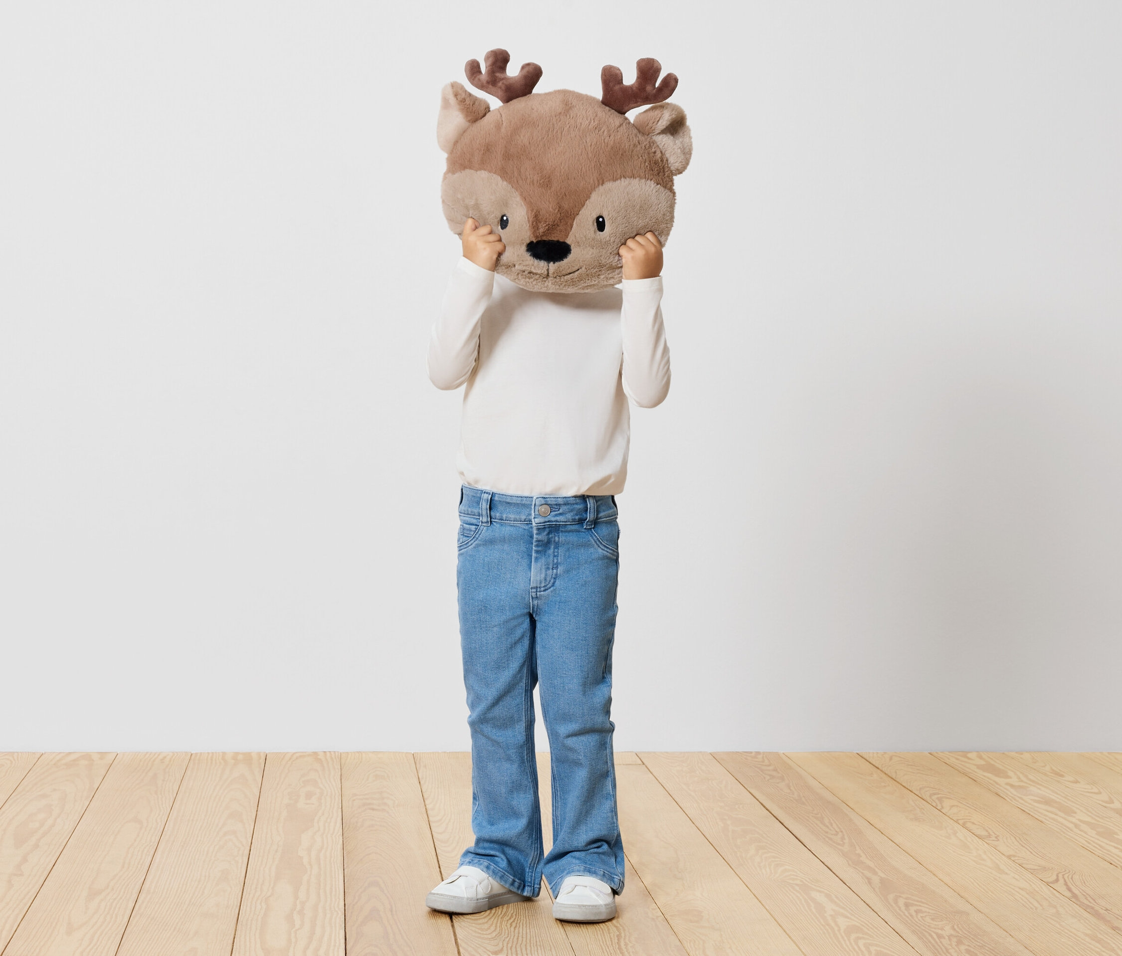 Dziecko trzyma pluszowego renifera przed twarzą, stojąc na drewnianej podłodze przed białą ścianą.