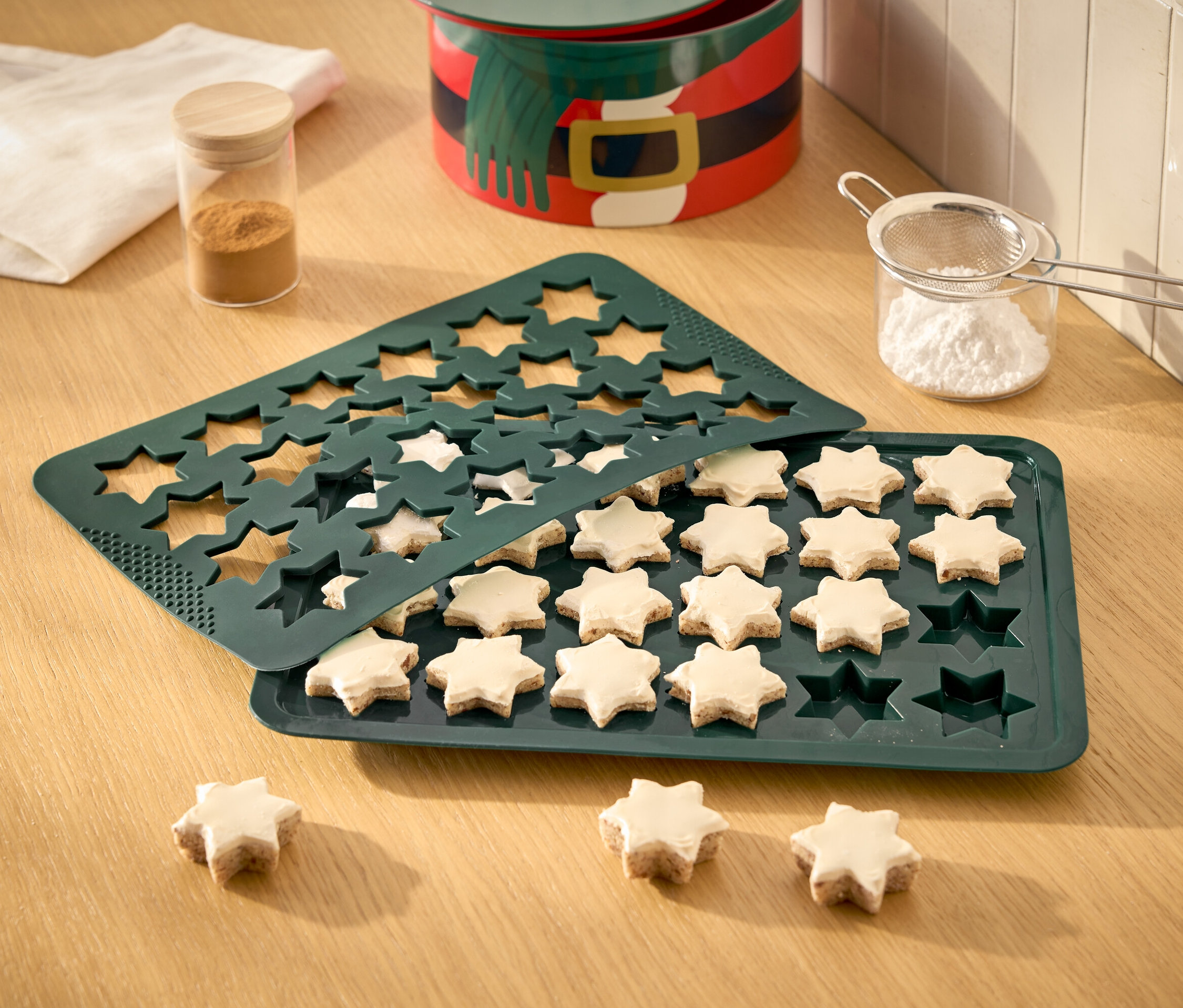 Gwiazdki w zielonej formie do pieczenia, niektóre już upieczone i udekorowane lukrem, przesiewacz do cukru pudru, puszka i ściereczka na drewnianym stole.