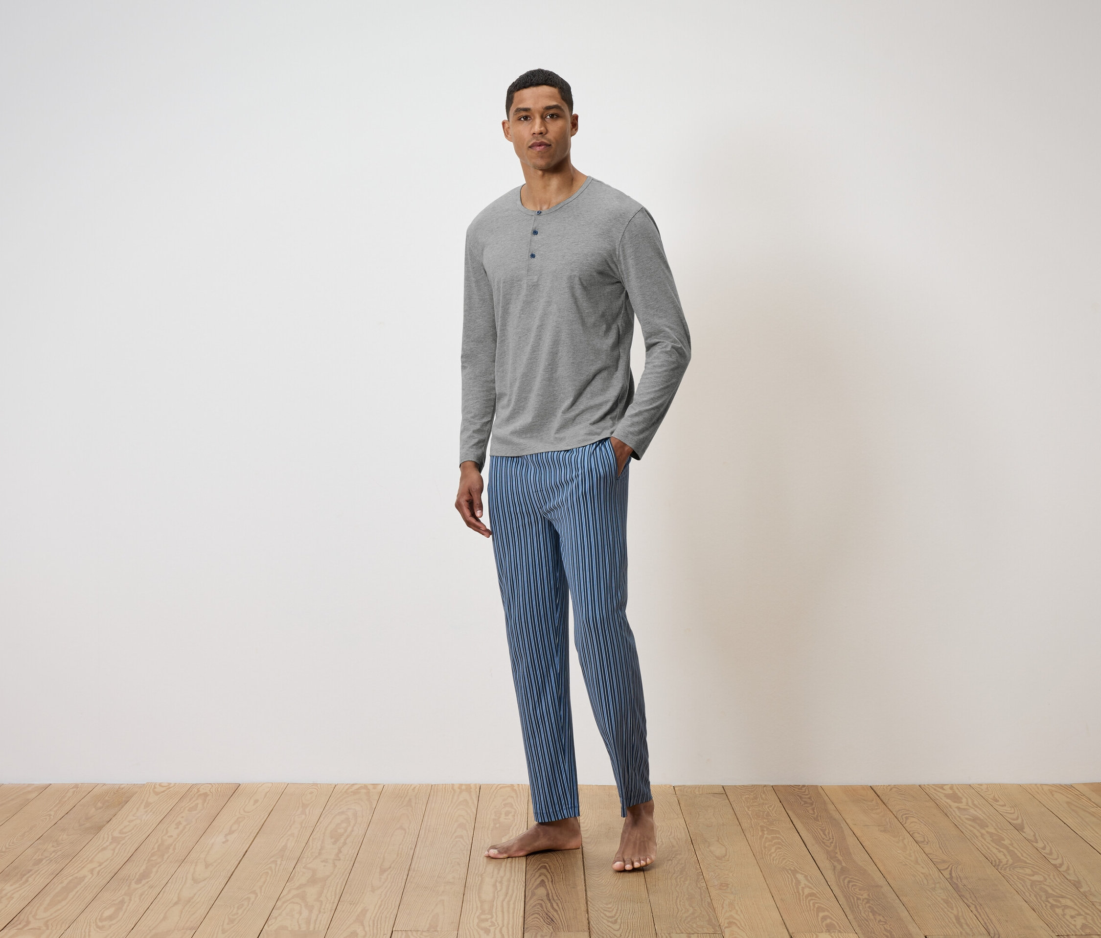 Mężczyzna stoi na drewnianej podłodze, ubrany w szarą koszulkę z długim rękawem i niebieskie spodnie w paski.