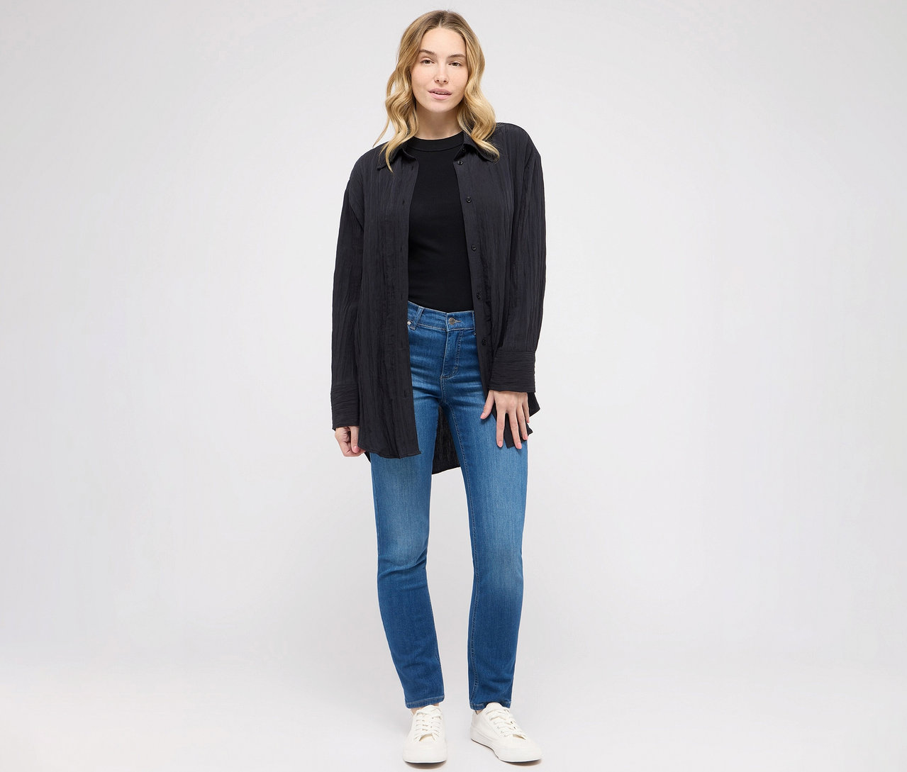 Kobieta pozuje w dżinsach Angels „Cici” Slim Fit, niebieskich, czarnym T-shircie i bluzce.
