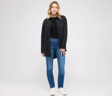 Kobieta pozuje w dżinsach Angels „Cici” Slim Fit, niebieskich, czarnym T-shircie i bluzce.