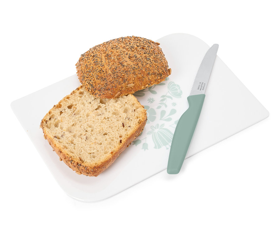 Na białej desce z kwiatowym wzorem leży kromka i cała bułka obok zielonego noża.