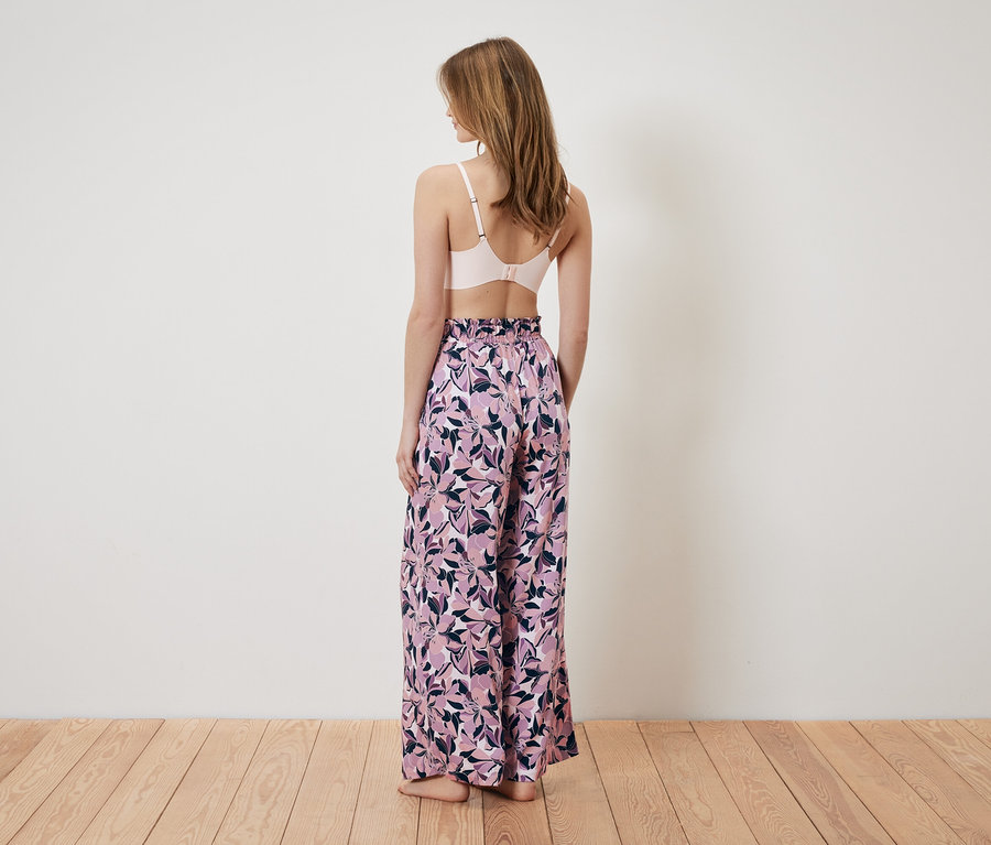 Kobieta tyłem do kamery ma na sobie różowy biustonosz i szerokie spodnie w kwiatowy wzór.