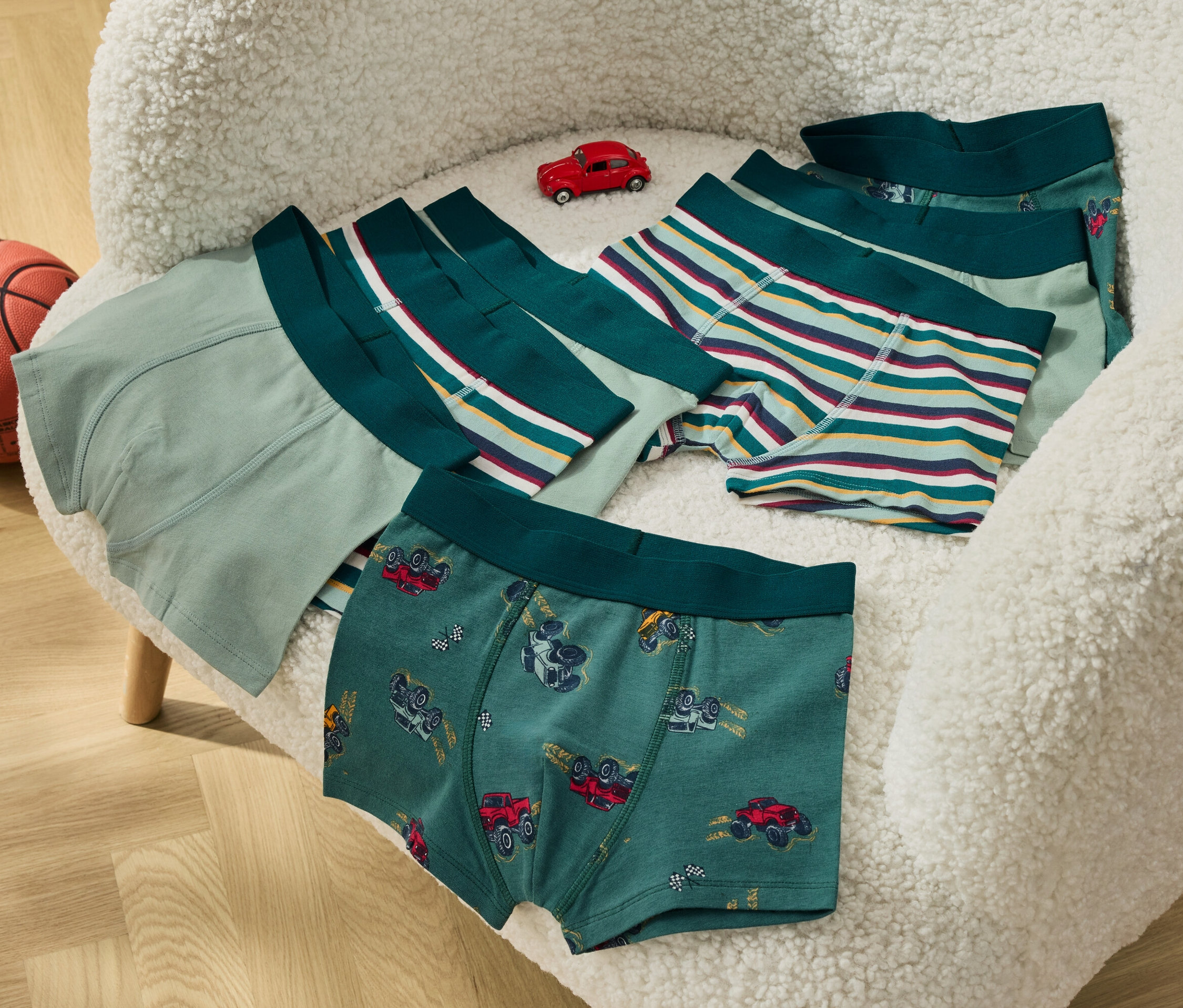 Na puszystym krześle leżą chłopięce bokserki i mały czerwony samochodzik, obok stoi piłka do koszykówki.