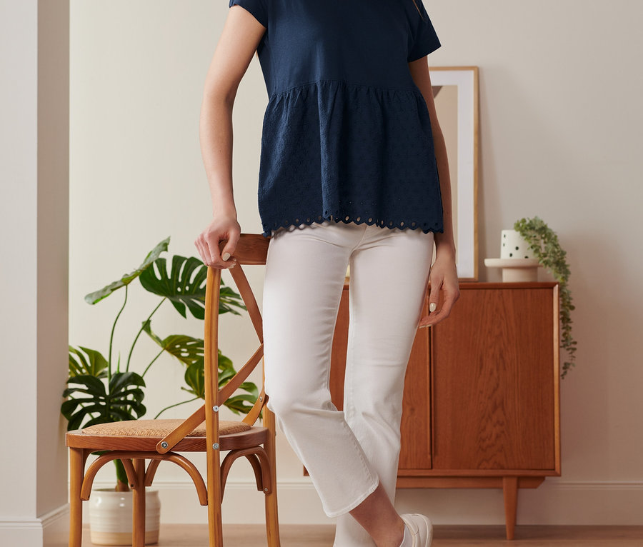 Kobieta oparta o krzesło ma na sobie granatową bluzkę z mieszanki materiałowej z haftem i białe spodnie.