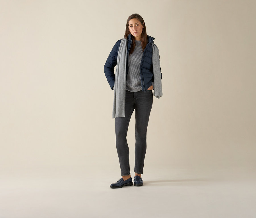 Kobieta stoi i ma na sobie spodnie dżinsowe o wąskim kroju – fason „Emma”, grey denim, szary sweter, granatową kurtkę i szary szalik.
