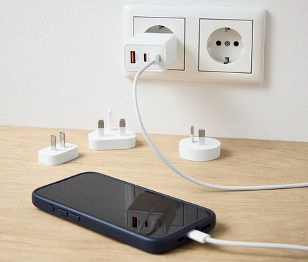 Biały adapter podróżny USB podłączony do gniazdka ładuje telefon komórkowy. Na stole leżą dodatkowe adaptery.
