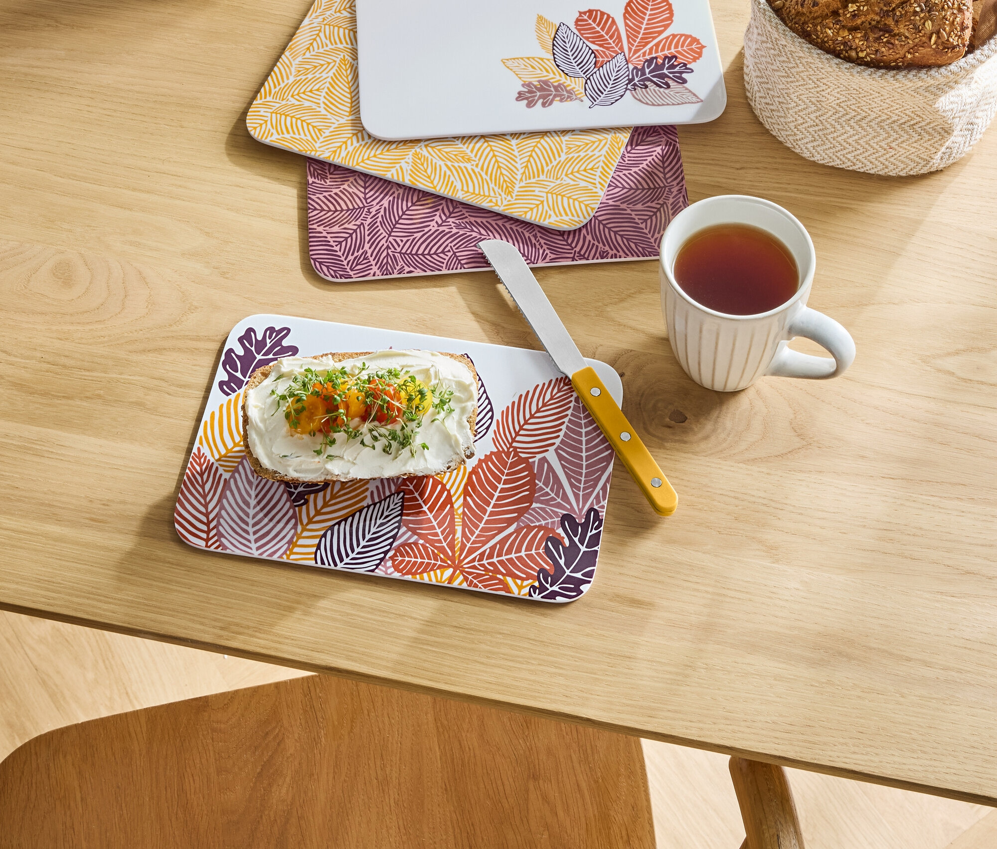Na stole leżą trzy deski z motywem liści, na jednej z nich chleb z serem i warzywami, obok stoi kubek z herbatą i koszyk z chlebem.
