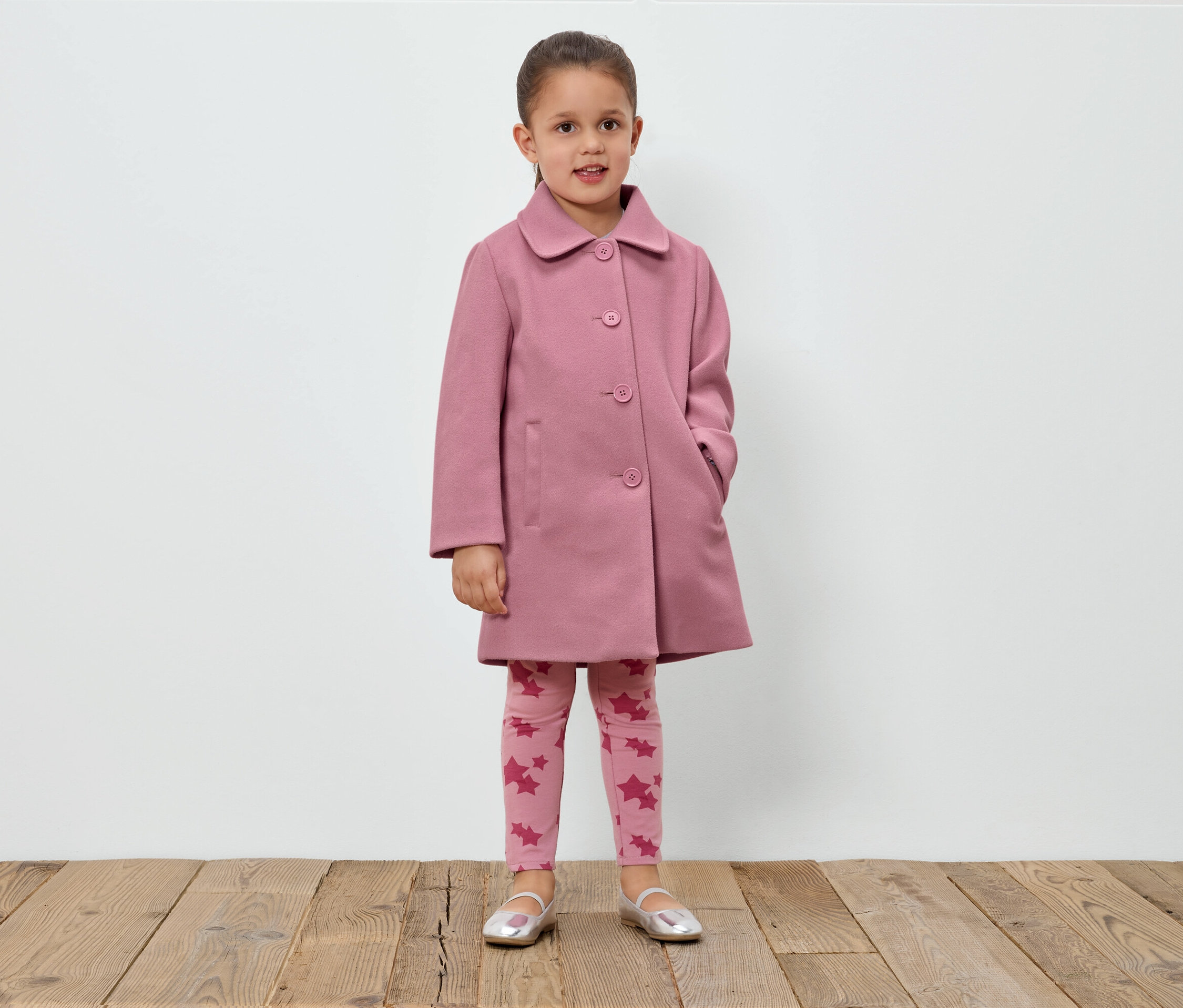Dziewczynka stoi na tle białej ściany, ubrana w różowy płaszcz, legginsy w gwiazdki i srebrne buty.
