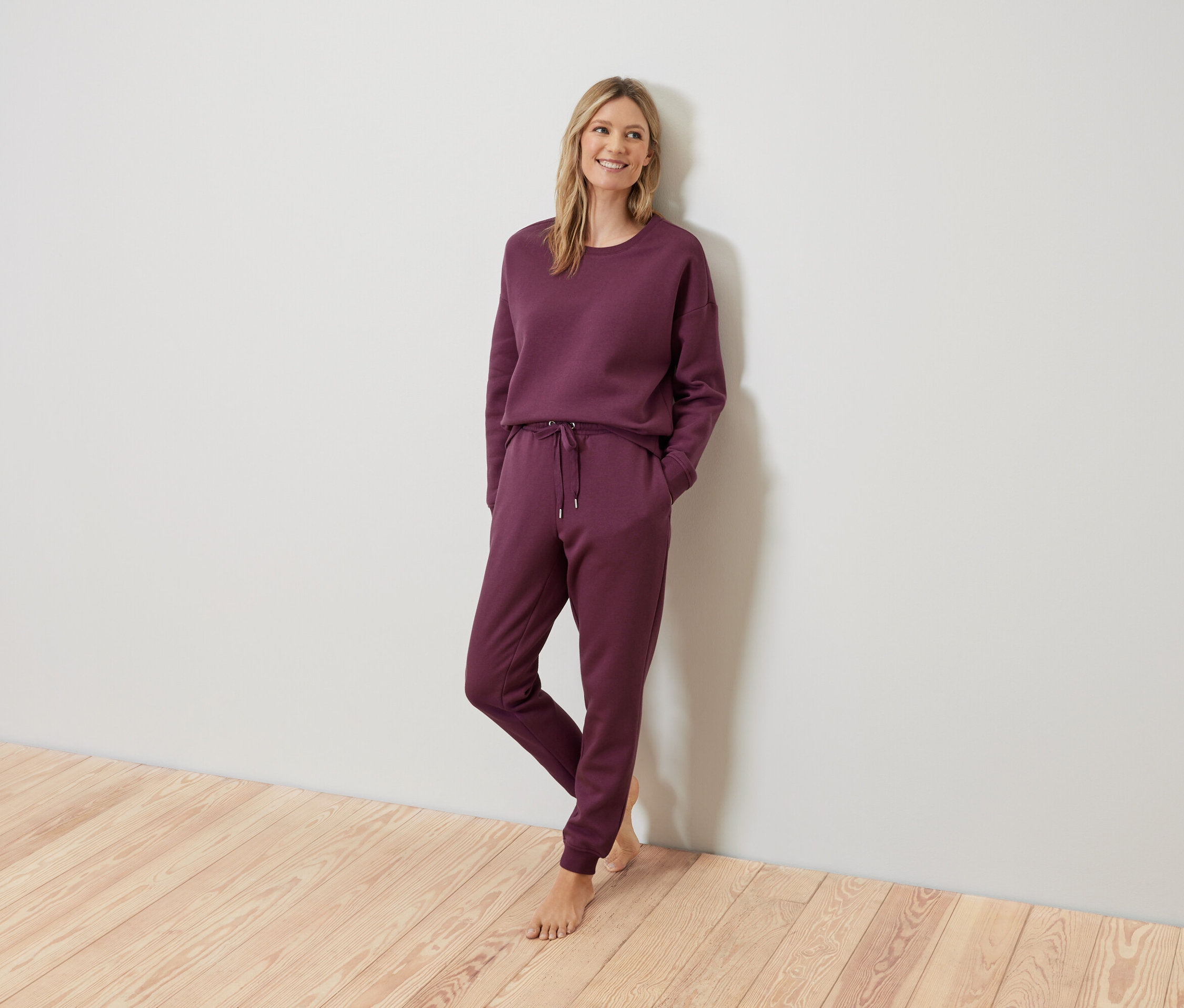 Blondynka pozuje w bordowym dresie przy ścianie.