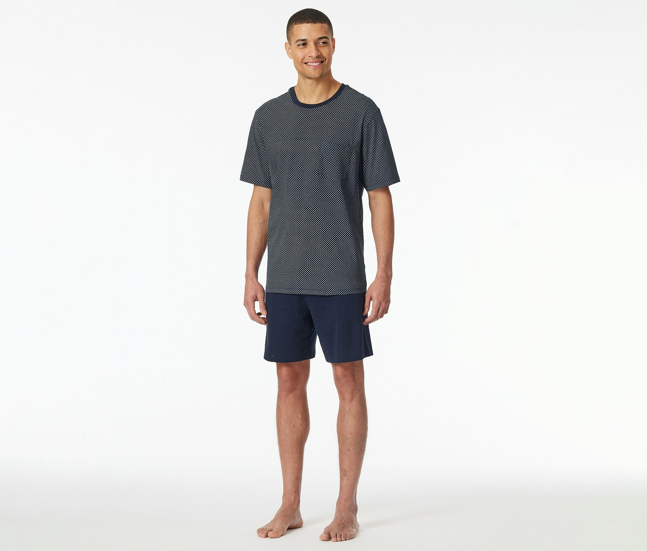 Mężczyzna stoi boso na białym tle w granatowym piżamie z krótkimi spodenkami SCHIESSER: koszulce w kropki z krótkim rękawem i szortach.