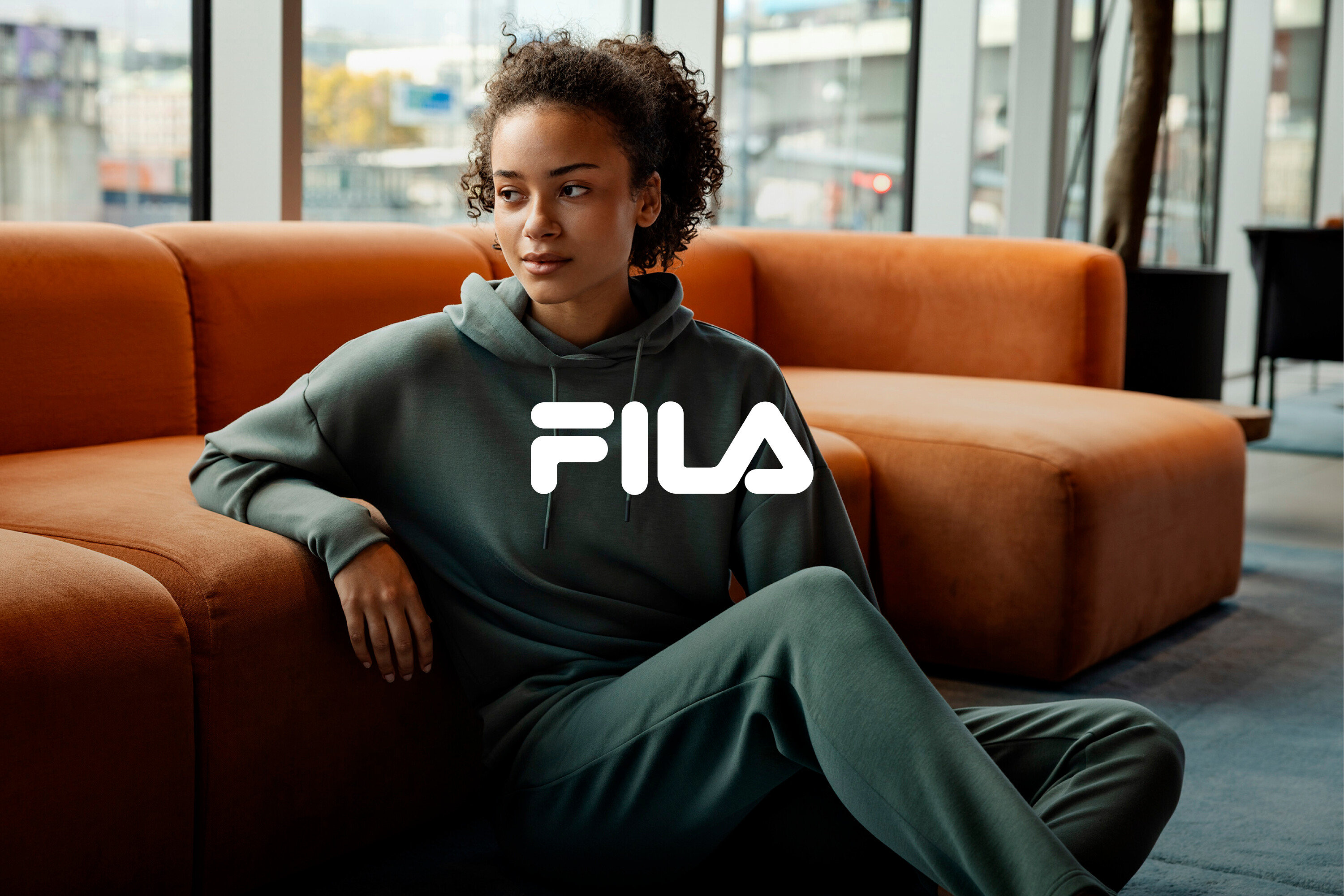 Kobieta siedzi na podłodze, oparta o pomarańczową sofę, ubrana w zielony dres marki Fila.