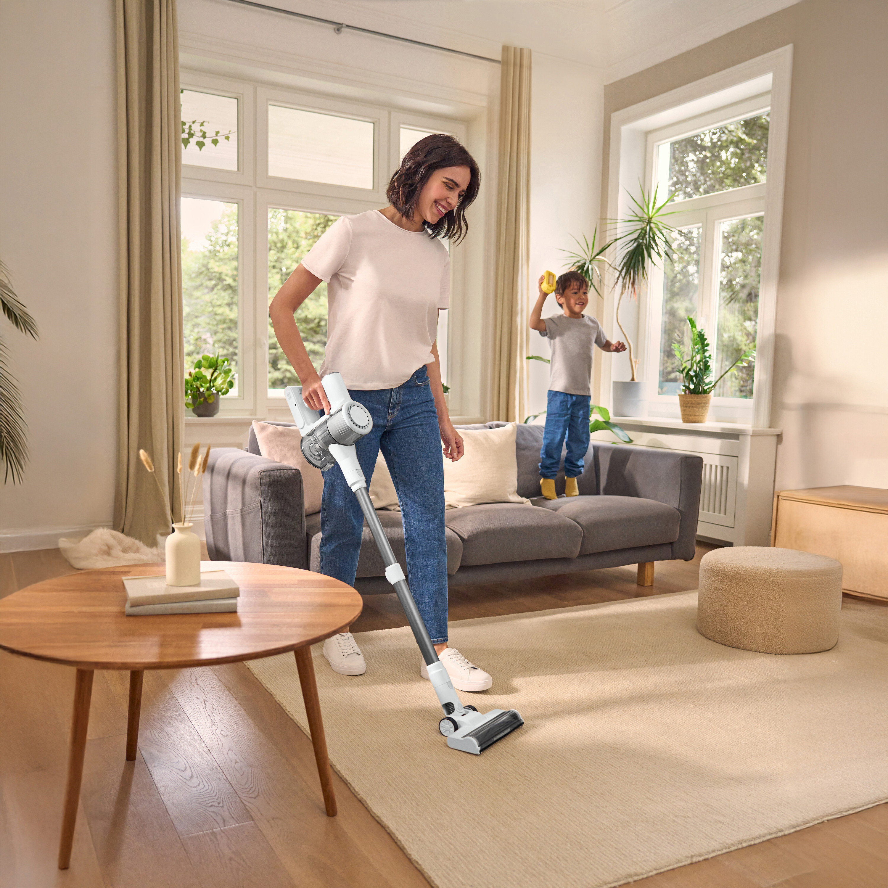 Kobieta odkurza dywan odkurzaczem akumulatorowym, dziecko stoi na sofie. Okrągły stół po lewej, okno w tle.