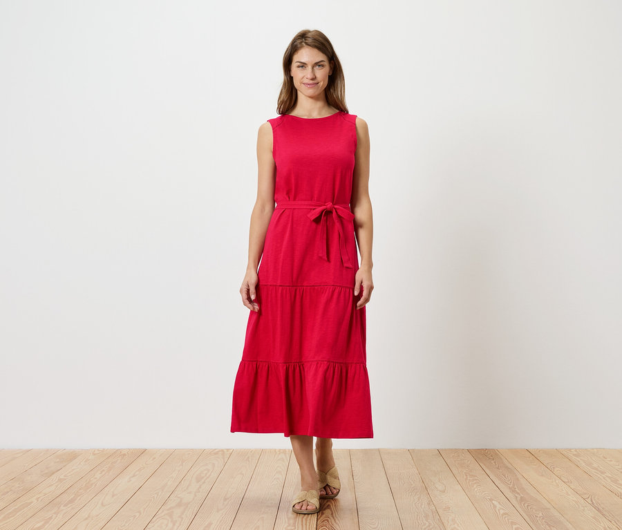 Kobieta stoi na tle białej ściany, ubrana w czerwoną, bezrękawną sukienkę z falbanami i paskiem oraz sandały.