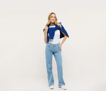 Kobieta pozuje w białym T-shircie i niebieskich dżinsach Angels „Liz” Wide Leg, z niebieskim swetrem na ramionach.