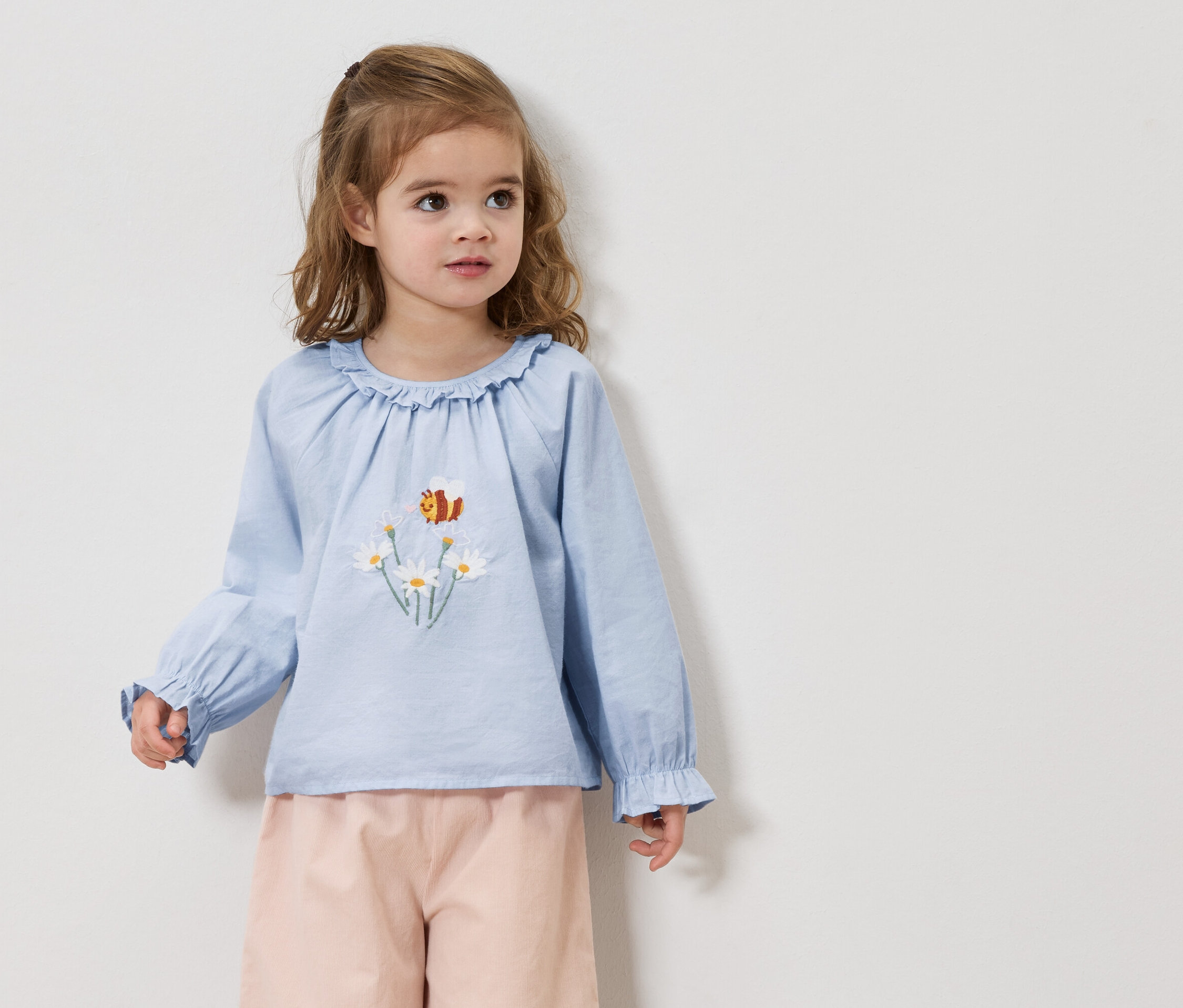 Dziewczynka z blond włosami ma na sobie jasnoniebieską bluzkę z haftowanymi stokrotkami i pszczołą oraz różowe spodnie.